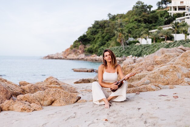 giovane donna caucasica calma felice con ukulele sulla spiaggia rocciosa tropicale al tramonto