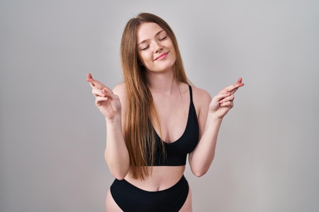 Giovane donna caucasica che indossa lingerie gesturing dito incrociato sorridendo con speranza e occhi chiusi fortuna e concetto superstizioso