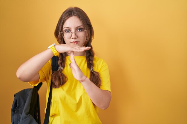 Giovane donna caucasica che indossa uno zaino da studente su sfondo giallo che fa un gesto di timeout con le mani, viso frustrato e serio