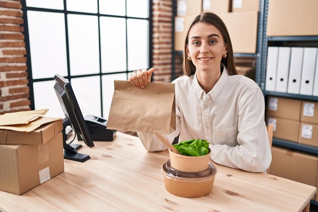 Giovane donna caucasica che lavora presso un e-commerce di piccole imprese che tiene cibo da asporto che sembra positivo e felice in piedi e sorride con un sorriso fiducioso che mostra i denti