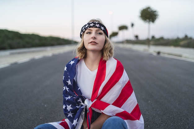 Giovane donna caucasica con capelli biondi corti e una bandiera americana seduta per strada