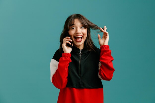 Giovane donna caucasica divertita che afferra i capelli guardando la fotocamera mentre parla al telefono isolato su sfondo blu