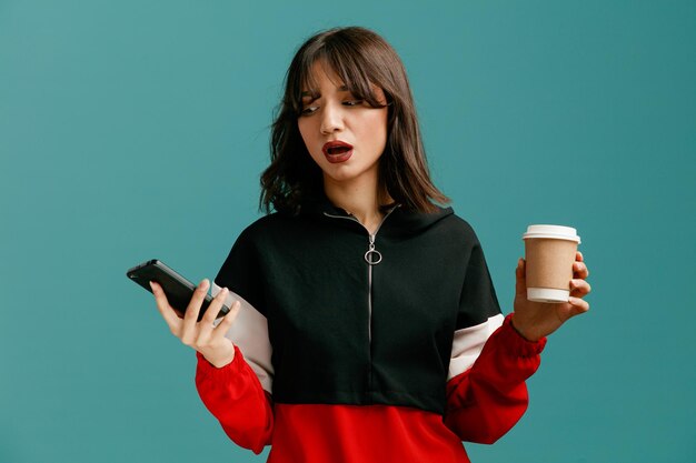 Giovane donna caucasica impressionata che tiene telefono cellulare e tazza di caffè di carta guardando il telefono cellulare isolato su sfondo blu