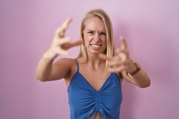 Giovane donna caucasica in piedi su sfondo rosa che urla frustrata dalla rabbia, mani che cercano di strangolare, urlando pazza