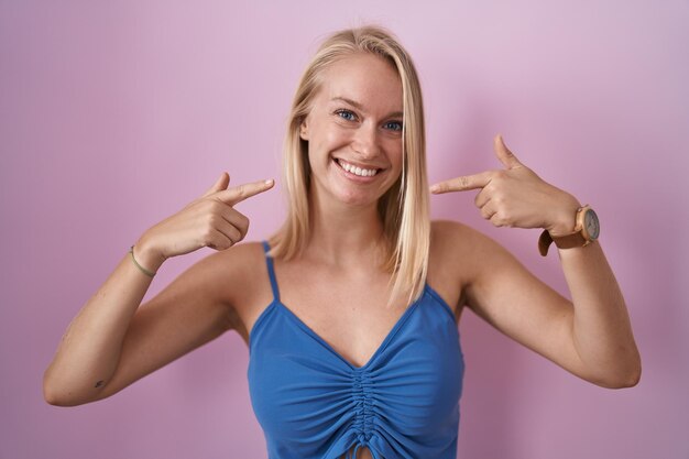 Giovane donna caucasica in piedi su sfondo rosa sorridente mostrando allegro e indicando con le dita i denti e la bocca. concetto di salute dentale.
