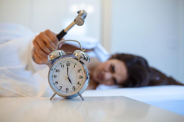 Giovane donna cerca di rompere la sveglia con un martello Distruggi l'orologio Ragazza sdraiata a letto spegnendo una sveglia con un martello al mattino alle 5 del mattino