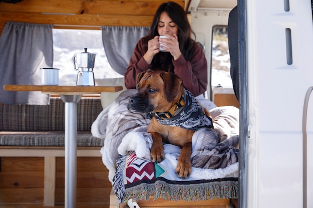 Giovane donna che accarezza il suo boxer e beve caffè dopo essersi svegliata in un camper durante il viaggio invernale