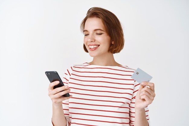 Giovane donna che acquista online con carta di credito in plastica e smartphone ragazza che legge lo schermo del telefono cellulare e sorride pagando su sfondo bianco di internet