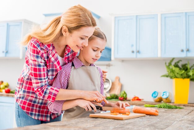 Giovane donna che aiuta sua figlia che taglia la carota con il coltello sulla tavola di legno