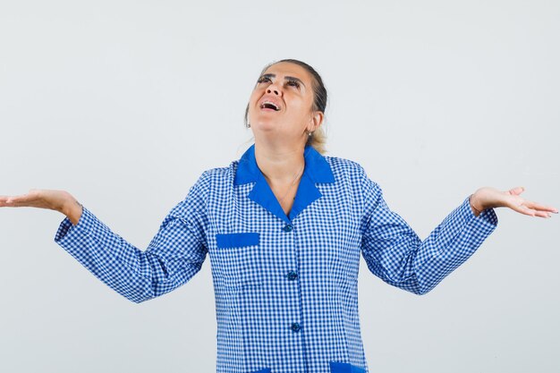 Giovane donna che allarga i palmi mentre riceve qualcosa mentre guarda sopra in camicia del pigiama a quadretti blu e sembra stupita. vista frontale.