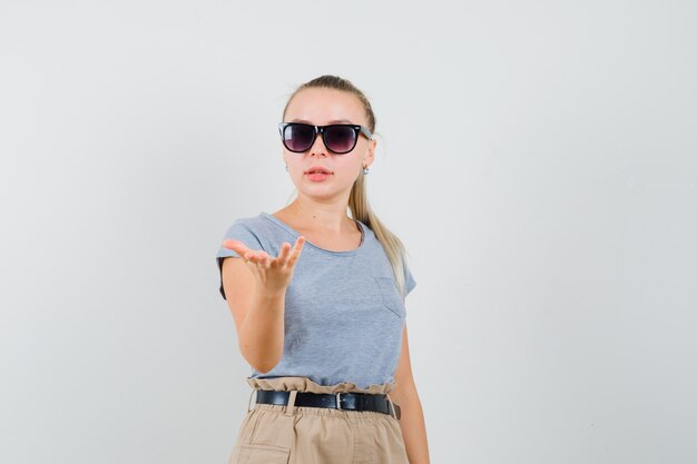 Giovane donna che allunga la mano nel gesto perplesso in t-shirt, pantaloni, vista frontale.