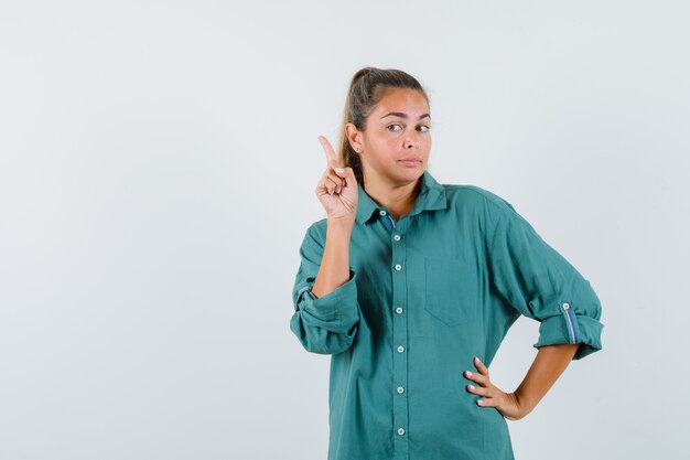 Giovane donna che alza il dito indice nel gesto di Eureka mentre tiene un'altra mano sulla vita in camicetta verde e sembra pensierosa