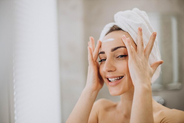 Giovane donna che applica la crema per il viso sul viso con un asciugamano sulla testa