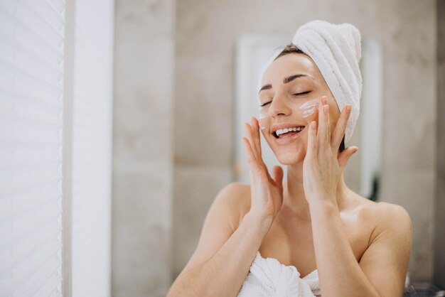 Giovane donna che applica la crema per il viso sul viso con un asciugamano sulla testa