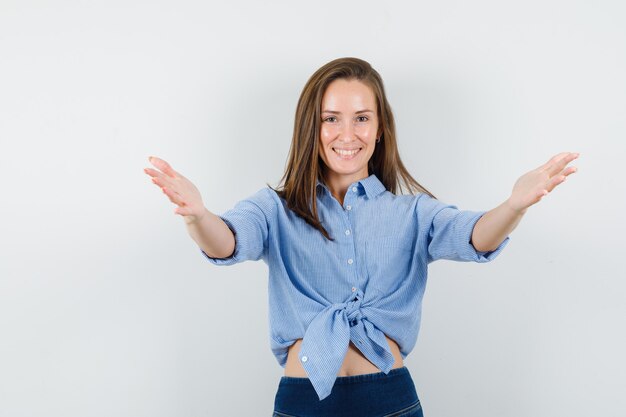 Giovane donna che apre le braccia per abbraccio in camicia blu, pantaloni e sembra contenta.