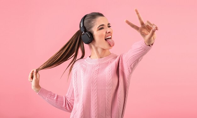 Giovane donna che ascolta la musica in cuffie senza fili che indossa un maglione rosa sorridente in posa sul rosa