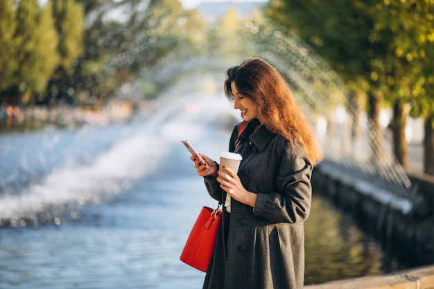 Giovane donna che beve caffè e che utilizza telefono nel parco