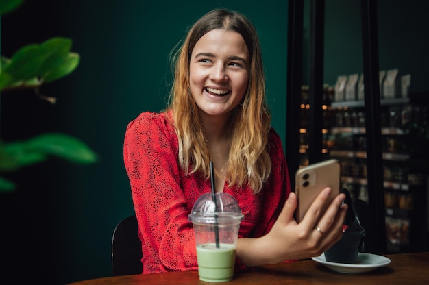 Giovane donna che beve il latte matcha del ghiaccio della bevanda verde nella caffetteria e utilizza lo smartphone