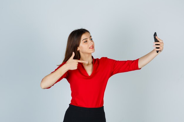 Giovane donna che cattura selfie mentre indica il lato destro in camicetta rossa