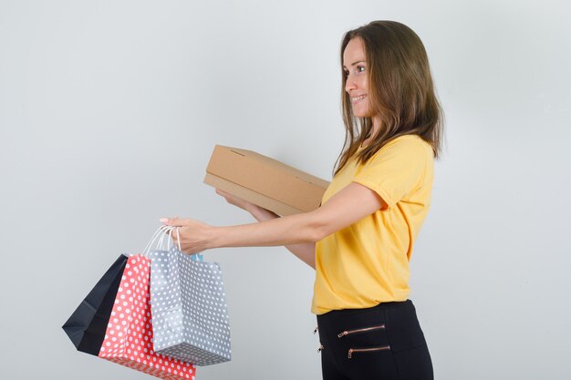 Giovane donna che consegna i sacchetti di carta con la scatola di cartone in maglietta gialla, pantaloni e che sembra contenta. .