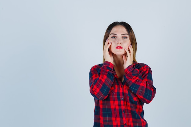 Giovane donna che controlla la pelle del viso toccando le guance in una camicia a quadri e sembra delicata, vista frontale.