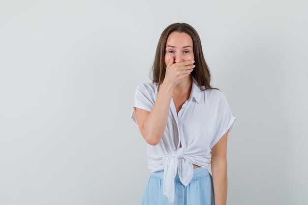 Giovane donna che copre la bocca con la mano in camicetta bianca e gonna azzurra e sembra allegra