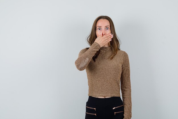 Giovane donna che copre la bocca con la mano in maglione dorato e pantaloni neri e sembra sorpresa