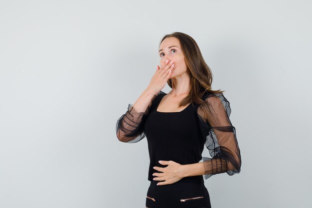Giovane donna che copre la bocca con una mano e che tiene un'altra mano sulla pancia in camicetta nera e pantaloni neri e sembra timida. vista frontale.