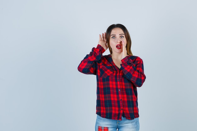 Giovane donna che dice il segreto tenendo una mano vicino alla bocca, tenendo l'altra dietro l'orecchio in camicia a quadri, jeans e sembra scioccata. vista frontale.