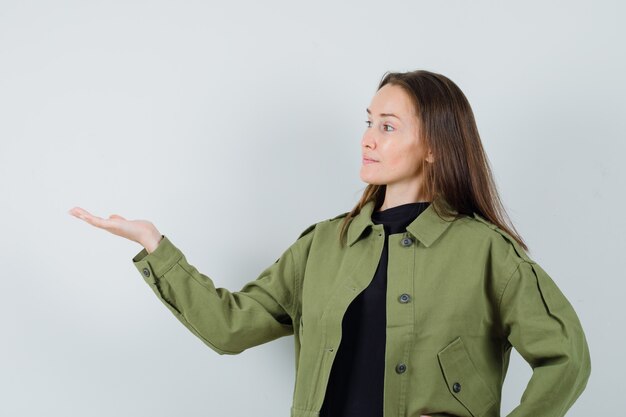 Giovane donna che diffonde il palmo da parte per mostrare qualcosa in giacca verde e guardare concentrato. vista frontale.