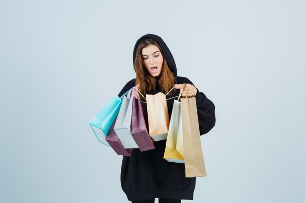 Giovane donna che esamina i pacchetti in felpa con cappuccio oversize, pantaloni e sembra sorpresa. vista frontale.