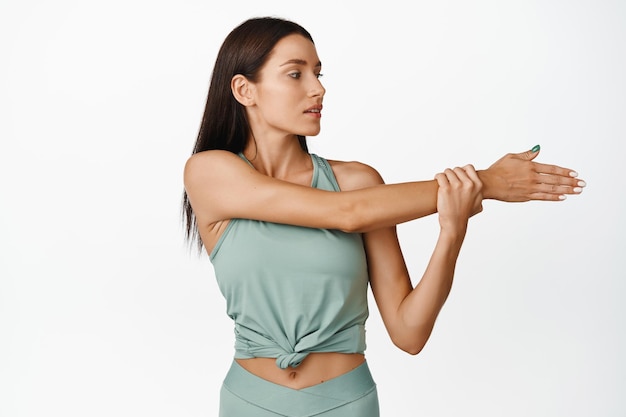 Giovane donna che fa allunga riscaldamento prima dell'allenamento mostrando esercizi di fitness in palestra in piedi su sfondo bianco