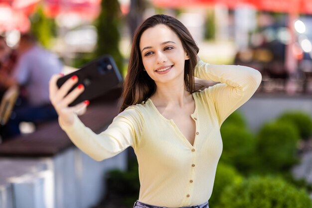 Giovane donna che fa selfie mentre beve caffè in città. Eccitata signora bianca in abito casual che si rilassa in cortile e si fotografa.