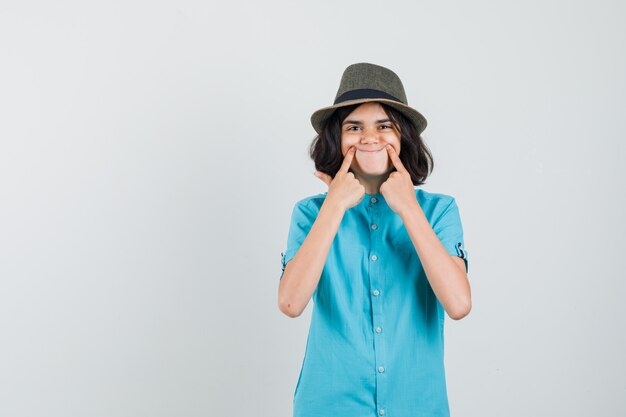 Giovane donna che fa un sorriso falso con le dita in camicia blu e sembra positivo