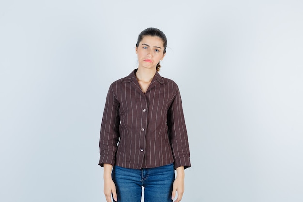 Giovane donna che fa una smorfia mentre posa in camicia a righe, jeans e sembra sconvolta, vista frontale.
