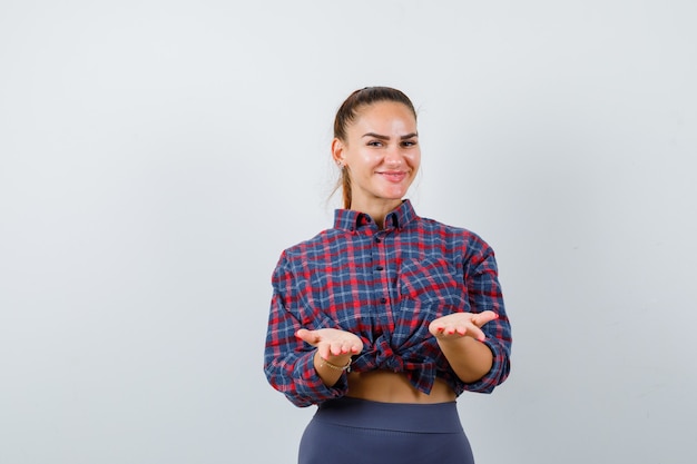 Giovane donna che finge di tenere qualcosa in camicia a scacchi, pantaloni e sembra felice, vista frontale.