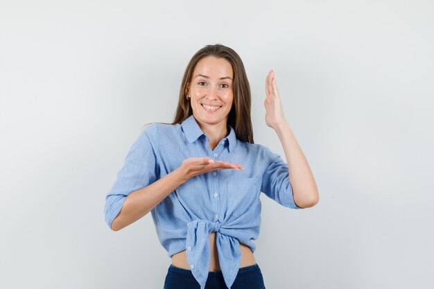 Giovane donna che gesturing con le mani alzate in camicia blu, pantaloni e sguardo allegro.