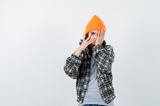 Giovane donna che guarda attraverso le dita con una camicia a scacchi con cappello arancione che sembra assonnata
