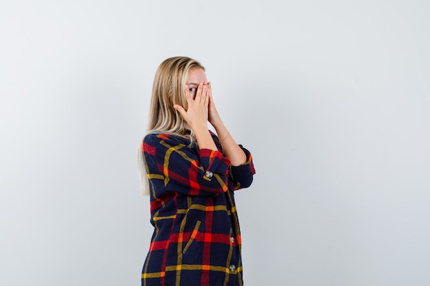 Giovane donna che guarda attraverso le dita mentre guarda le mani sul viso in camicia a quadri e sembra carina, vista frontale.