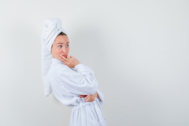 Giovane donna che guarda lontano con la mano sul mento in accappatoio bianco, asciugamano e guardando curioso.