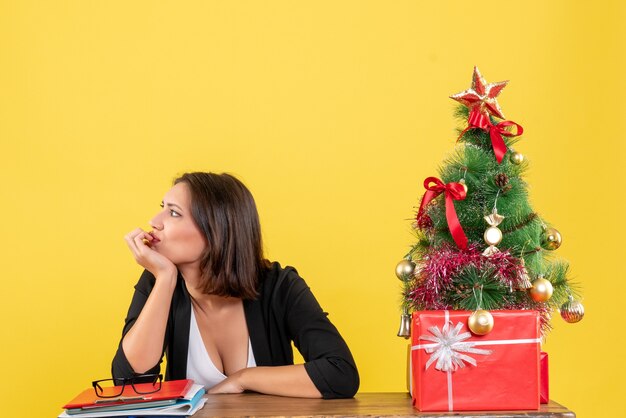 Giovane donna che guarda qualcosa seduto a un tavolo vicino all'albero di Natale decorato in ufficio su giallo