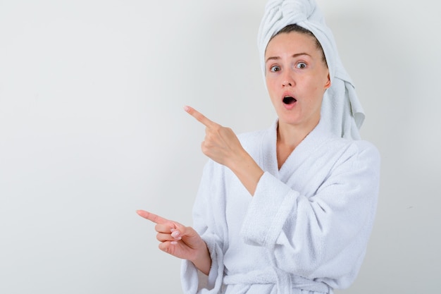 Giovane donna che indica da parte in accappatoio bianco, asciugamano e guardando stupito, vista frontale.
