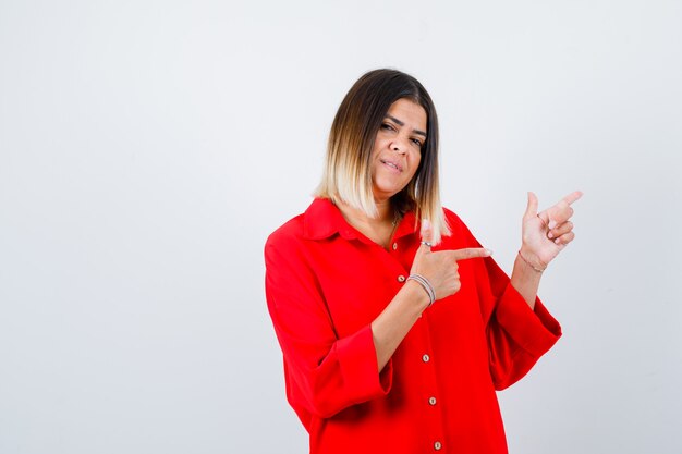 Giovane donna che indica da parte in camicia rossa oversize e sembra soddisfatta, vista frontale.