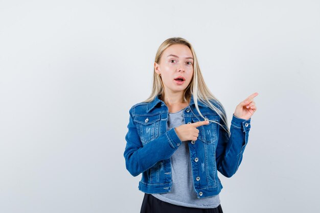 Giovane donna che indica da parte in maglietta, giacca di jeans, gonna e sembra stupita