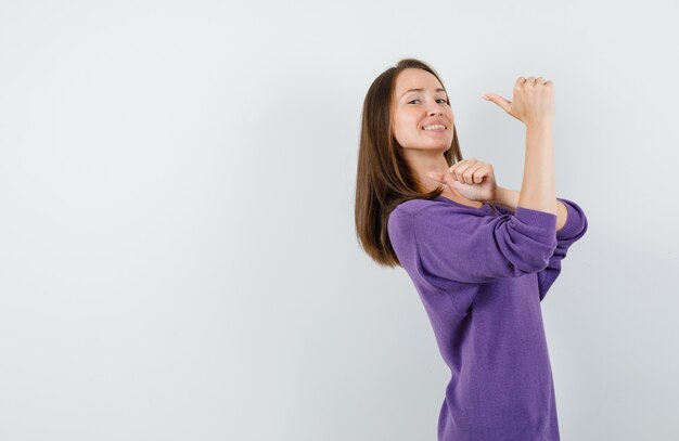 Giovane donna che indica i pollici indietro in camicia viola e guardando divertito, vista frontale.