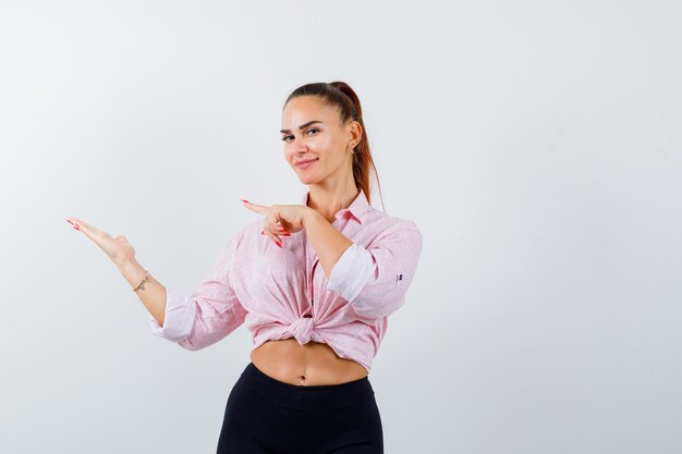 Giovane donna che indica il suo palmo aperto da parte in camicia, pantaloni e sembra sicura. vista frontale.