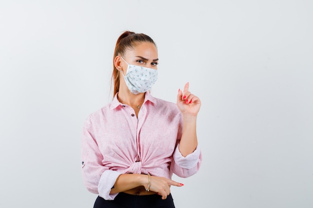 Giovane donna che indica in alto ea destra in camicia, maschera e sguardo pensieroso. vista frontale.