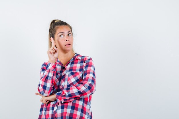 Giovane donna che indica in alto in camicia a quadri e sembra intelligente, vista frontale.