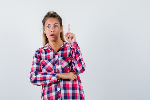 Giovane donna che indica in su in camicia a quadri e sembra scioccata. vista frontale.