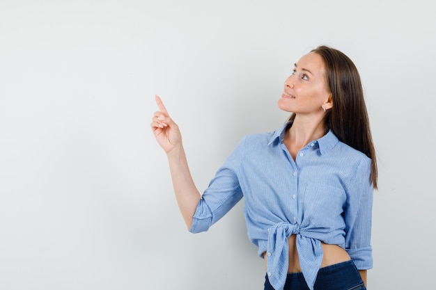 Giovane donna che indica in su in camicia blu, pantaloni e sembra allegra